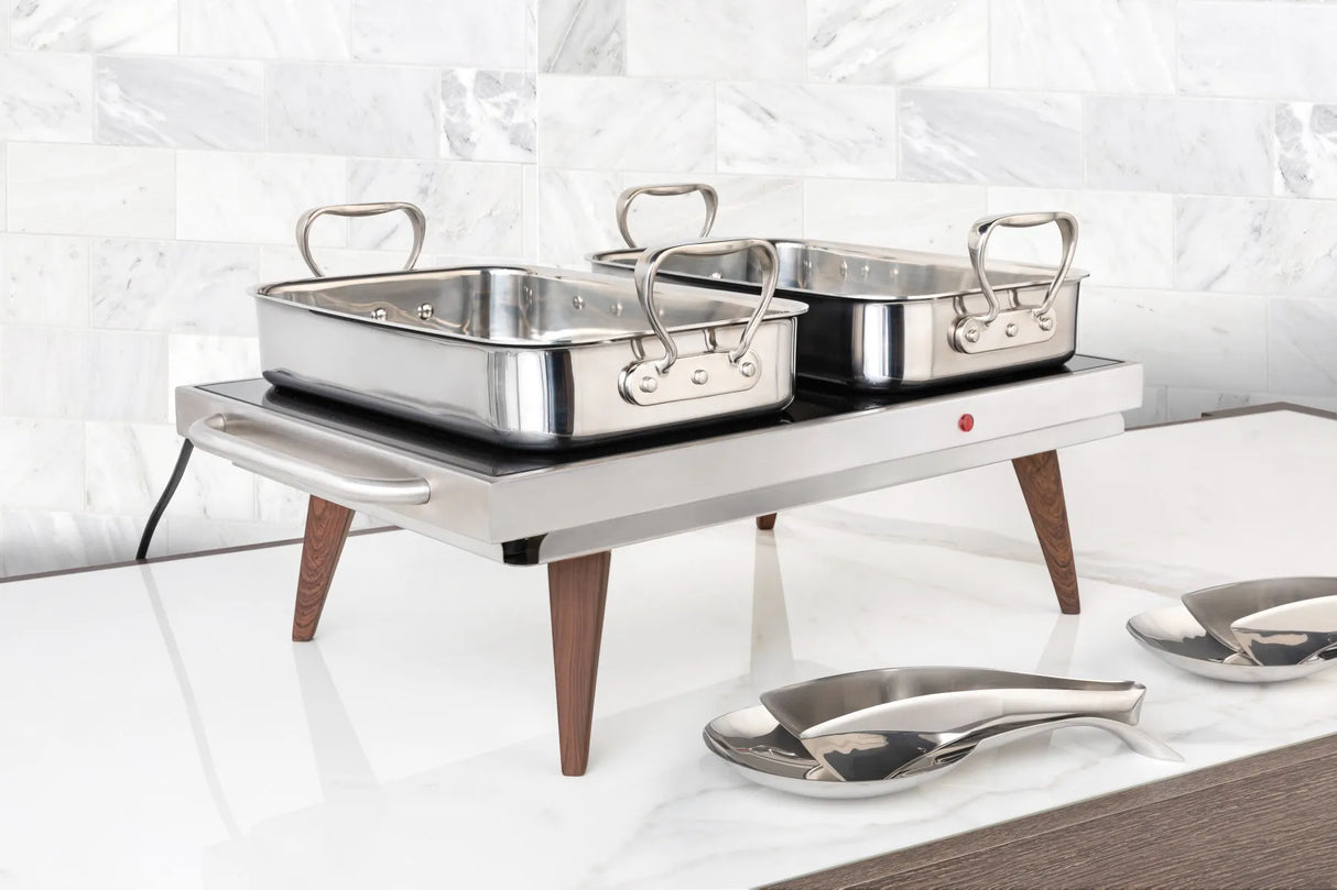 A stainless steel buffet roaster warming tray with wooden legs holds two 3 qt rectangular metal chafing dishes with handles. The tray sits on a white countertop near matching serving utensils and a white marble tile backsplash.