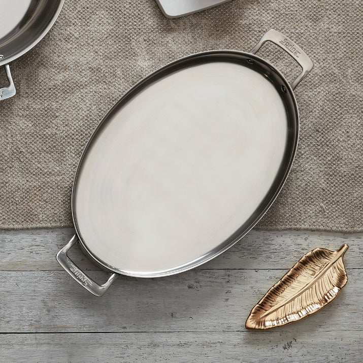 An empty, oval stainless steel serving tray with handles rests on a burlap cloth, perfect for buffet sauté presentations. Nearby, a metallic gold, leaf-shaped dish sits on a rustic wooden surface, with another metal dish peeking from the top.