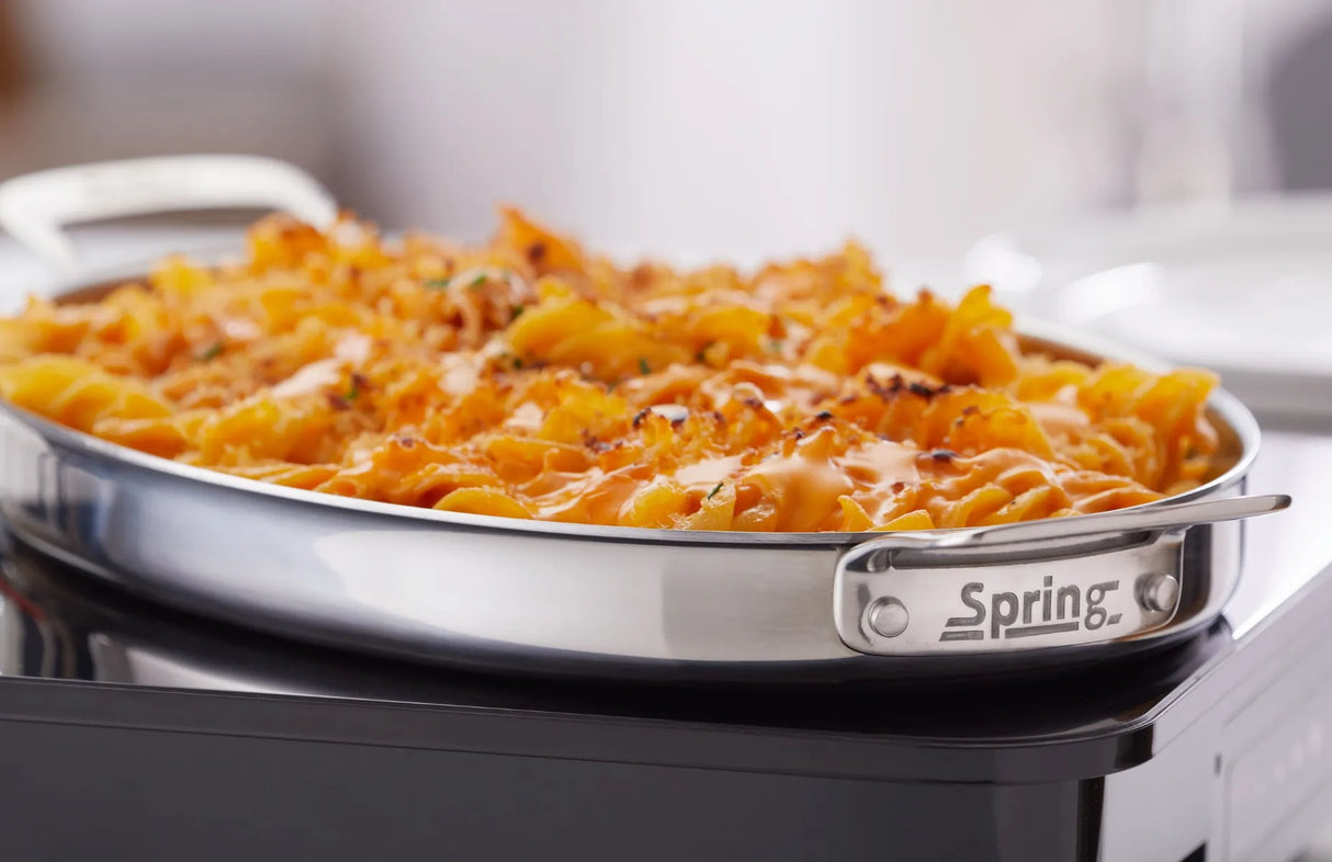 A 1-1/2 qt. oval stainless steel pan labeled “Spring” contains baked pasta topped with melted orange cheese and sprinkled herbs, sitting on a black stovetop. The crispy, golden edges make it perfect for a buffet sauté presentation.