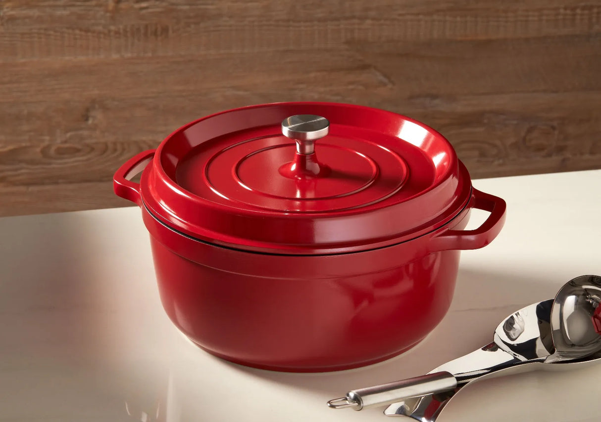 A shiny red Ironlite Casserole Dutch oven with a lid and metal knob sits on a glossy white countertop. The Dutch oven, perfect for induction cooking, has two side handles. A metal ladle and serving spoon rest nearby against a wood-paneled wall.