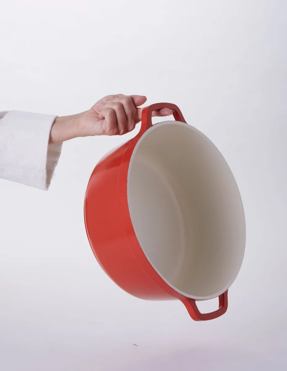 A person wearing a white sleeve holds a large, empty, round red Ironlite Casserole pot with two handles against a plain white background. The pot is tilted to display its light, non-stick casserole interior.