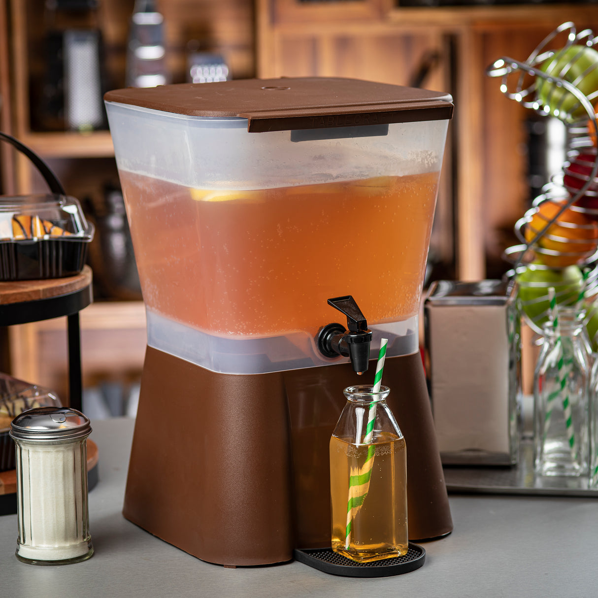 A 3 gallon beverage dispenser filled with iced tea stands on a counter. A glass bottle with a green-and-yellow striped straw is being filled from the spout. Sugar shaker, glasses, and kitchen shelves are visible in the background.
