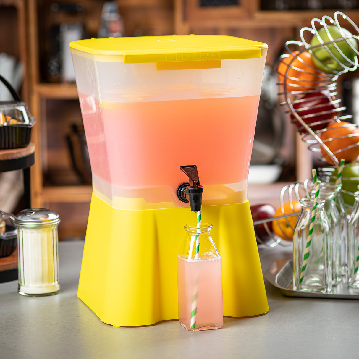 A 3-gallon beverage dispenser filled with pink lemonade stands on a counter. A glass bottle with a green-striped straw is positioned under the spout, half full of lemonade. Nearby are glass bottles, fruit in a spiral rack, and a sugar dispenser.
