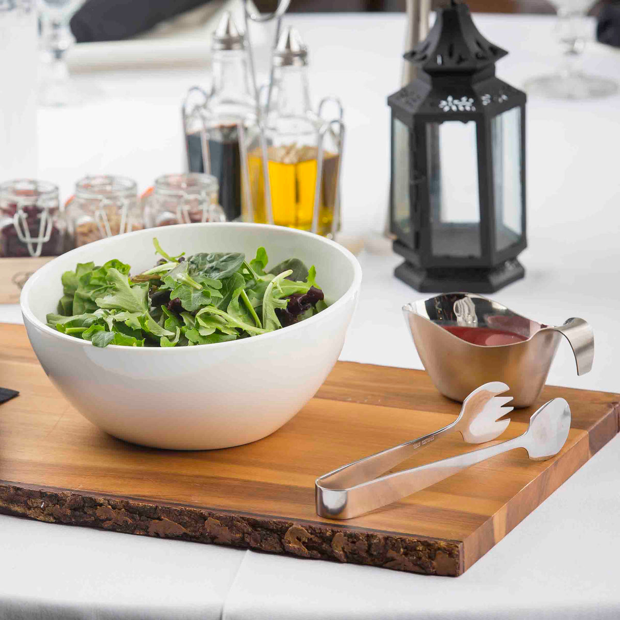 A white bowl of mixed leafy greens sits on a wooden board with stainless steel salad tongs, next to a small gravy boat with dressing. In the background are oil and vinegar bottles, jars, and a black lantern on a white tablecloth.