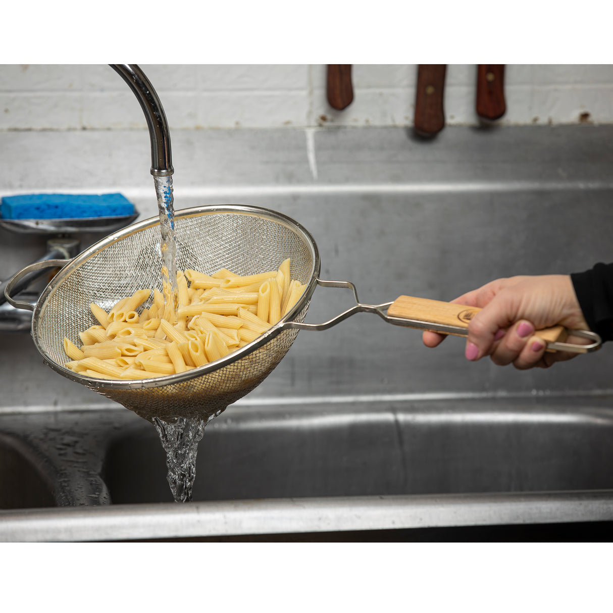 A hand holds a wooden-handle double medium mesh strainer filled with cooked penne pasta under a running faucet in a stainless steel kitchen sink, rinsing the pasta. Knives hang on a magnetic strip above the sink, and a blue sponge sits nearby.