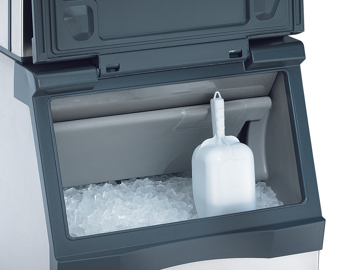 A close-up of a top-mounted ice maker shows an NSF-certified ice bin filled with small, clear ice cubes. A white plastic scoop rests upright on the ice inside the bin, and part of the machine’s dark gray exterior is visible.
