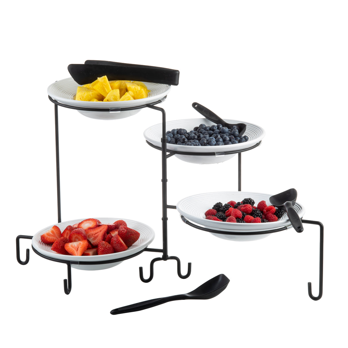 A three-tiered black powder coated metal stand holds four white bowls filled with cut pineapple, blueberries, strawberries, and mixed berries. Each bowl has a black serving tong or spoon. The stand is set on a white surface.