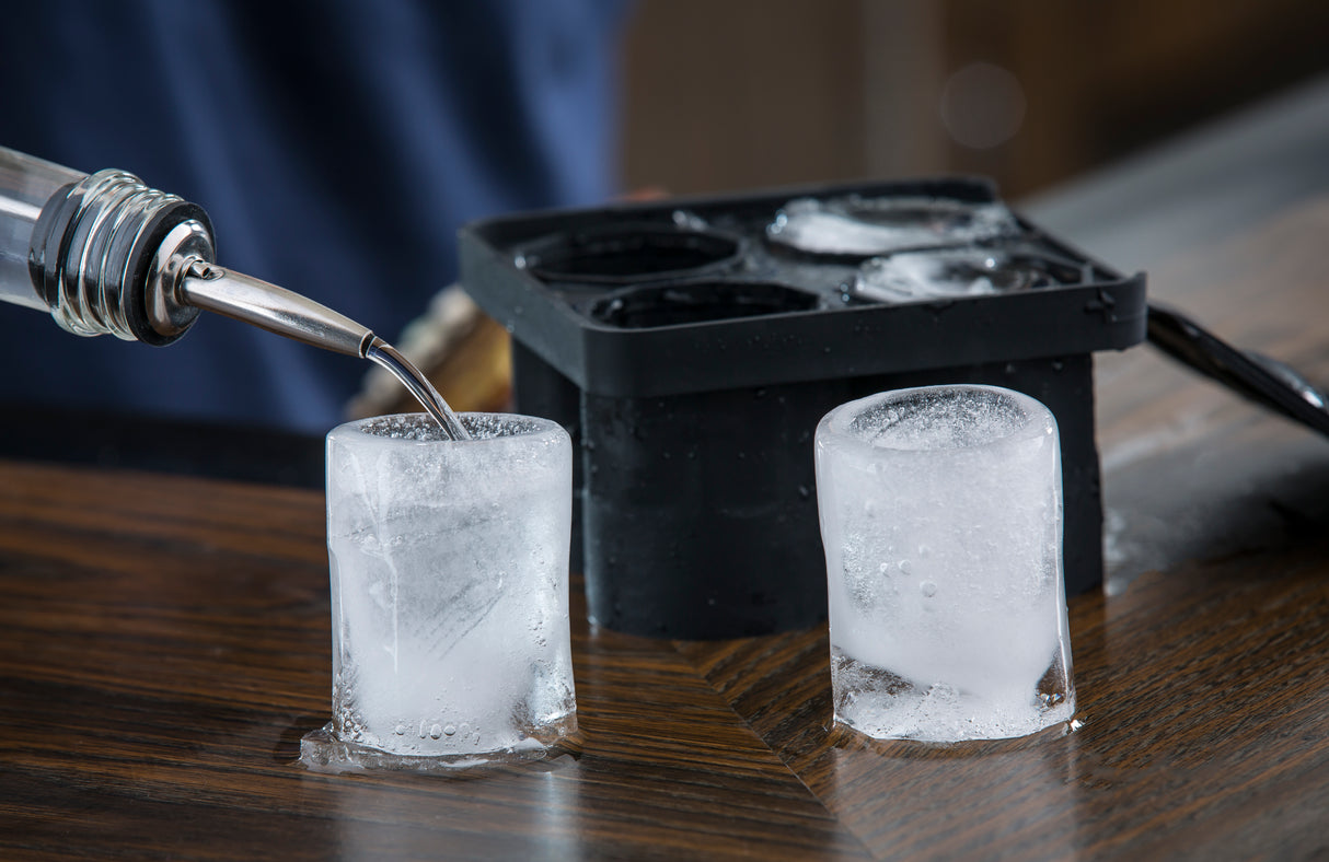 A close-up of two cylindrical silicone shot glasses on a wooden table, one being filled with clear liquid. In the background, a black shot glass ice tray is visible, along with a blurred dark blue shirt and brown surface.