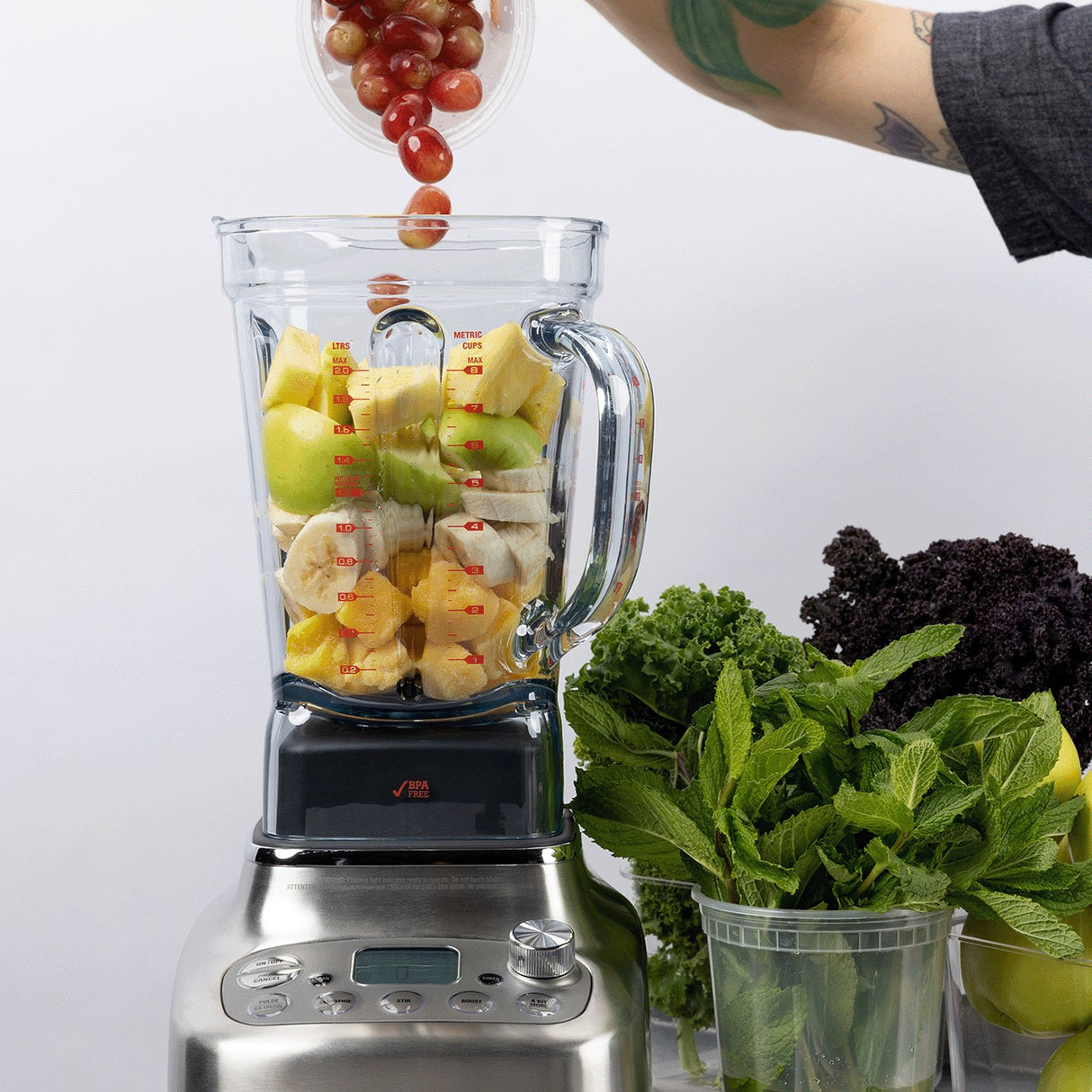 A person adds red grapes to a Breville Commercial Super Q Pro Blender filled with sliced bananas, green apples, and pineapple. Fresh greens like kale and mint sit nearby. The persons tattooed arm is visible by the 68 oz capacity stainless steel base.
