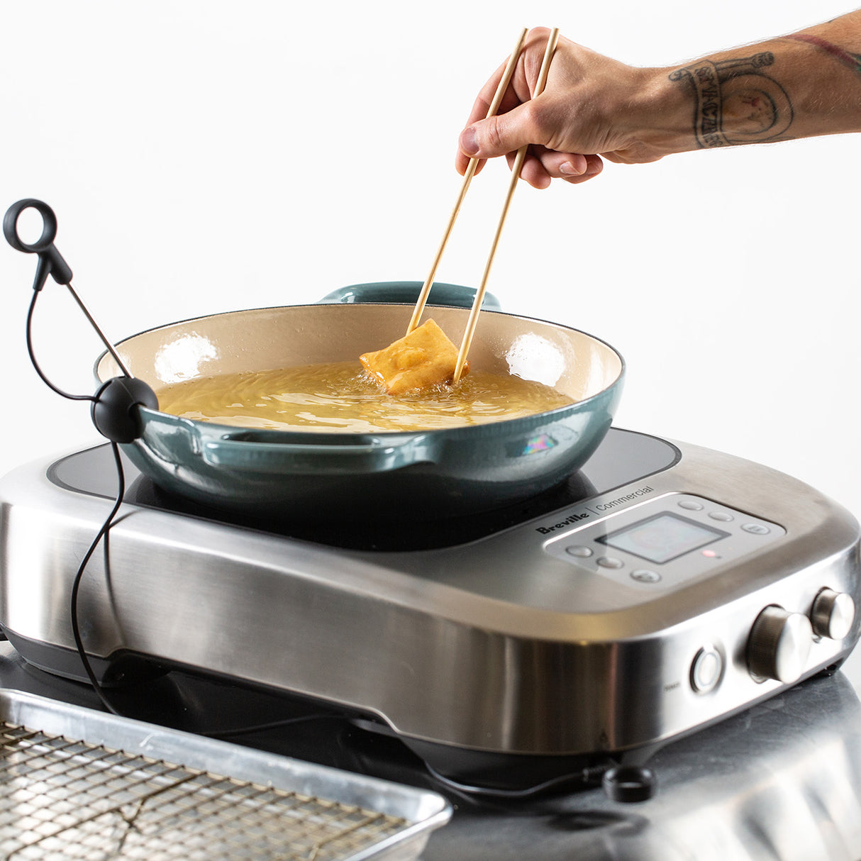 A hand holds chopsticks to fry food in a deep, light green pan filled with oil, set on a commercial induction stove for precise temperature control. A thermometer is clipped to the pan, and a cooling rack sits nearby against a minimalistic white background.