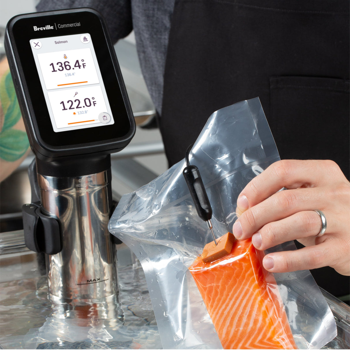 A person uses a Breville Commercial sous vide immersion circulator to cook vacuum-sealed salmon in water. They insert a temperature probe into the sealed bag; the display reads 136.4°F (water) and 122.0°F (salmon). The person wears a black apron.