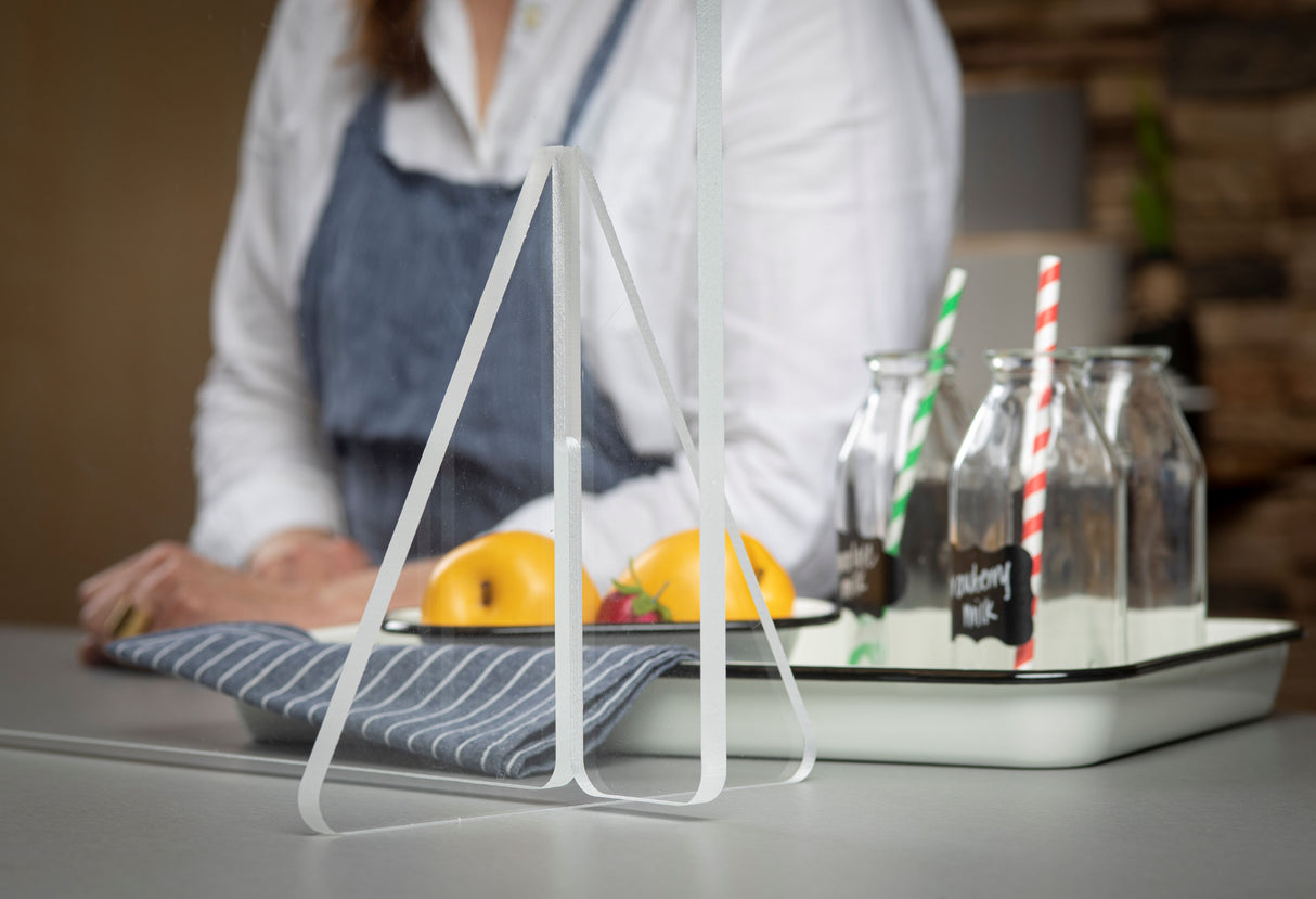 A woman in a white shirt and blue apron stands behind a counter. On the counter are glass bottles with chalk label tags, striped paper straws, a tray of yellow apples, and a striped cloth napkin. A portable countertop acrylic safety shield is in the foreground.