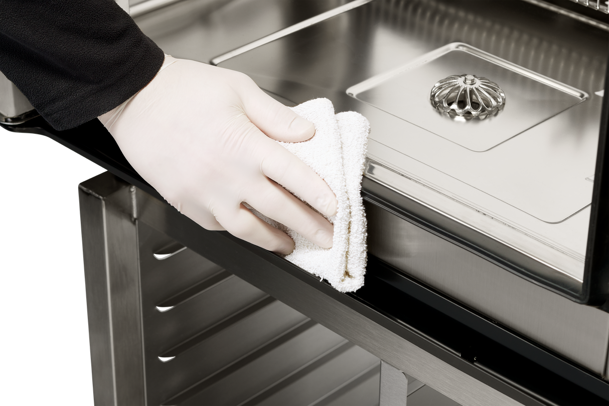 A person wearing a white glove wipes the edge of a Vulcan TCM-101E touch combi oven food service cart with a white cloth. The cart's shiny, reflective surface holds metal trays, one with a decorative lid, against a plain, uncluttered background.