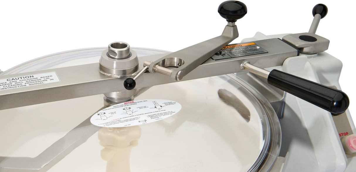 Close-up of a Hobart HCM450-20 Cutter Mixer with a large round transparent lid secured by a metal arm and black handles. Caution and instruction labels are visible, hinting at its sealing or pressurizing mechanism; includes Strainer Basket.