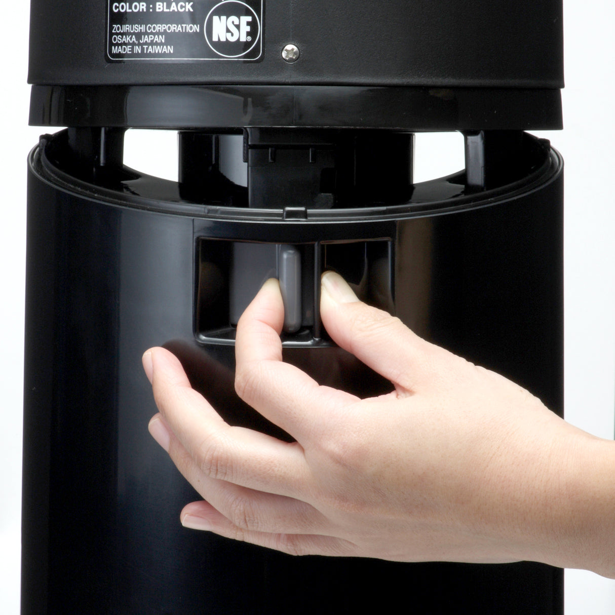 A close-up of a hand pressing a gray lever on a black insulated beverage dispenser. The background is plain white, and part of a product label with the text NSF and manufacturing details is visible at the top.