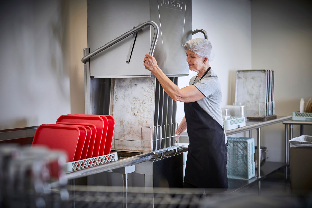 An older woman in a hairnet, grey shirt, and black apron operates a ventless dishwashing machine in a commercial kitchen, lifting its door. Clean red trays are stacked on stainless steel counters, highlighting the orderly and bright setting.