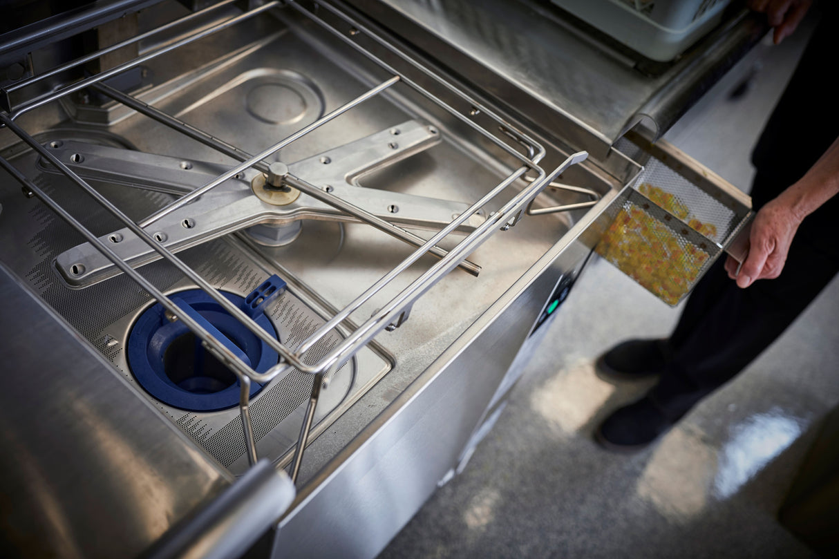 Close-up of an open ventless dishwashing machine with metal racks, a spinning spray arm, and a blue filter inside. A person’s hand pulls out a mesh filter tray filled with food scraps. The clean, metallic surfaces highlight high temp sanitizing.