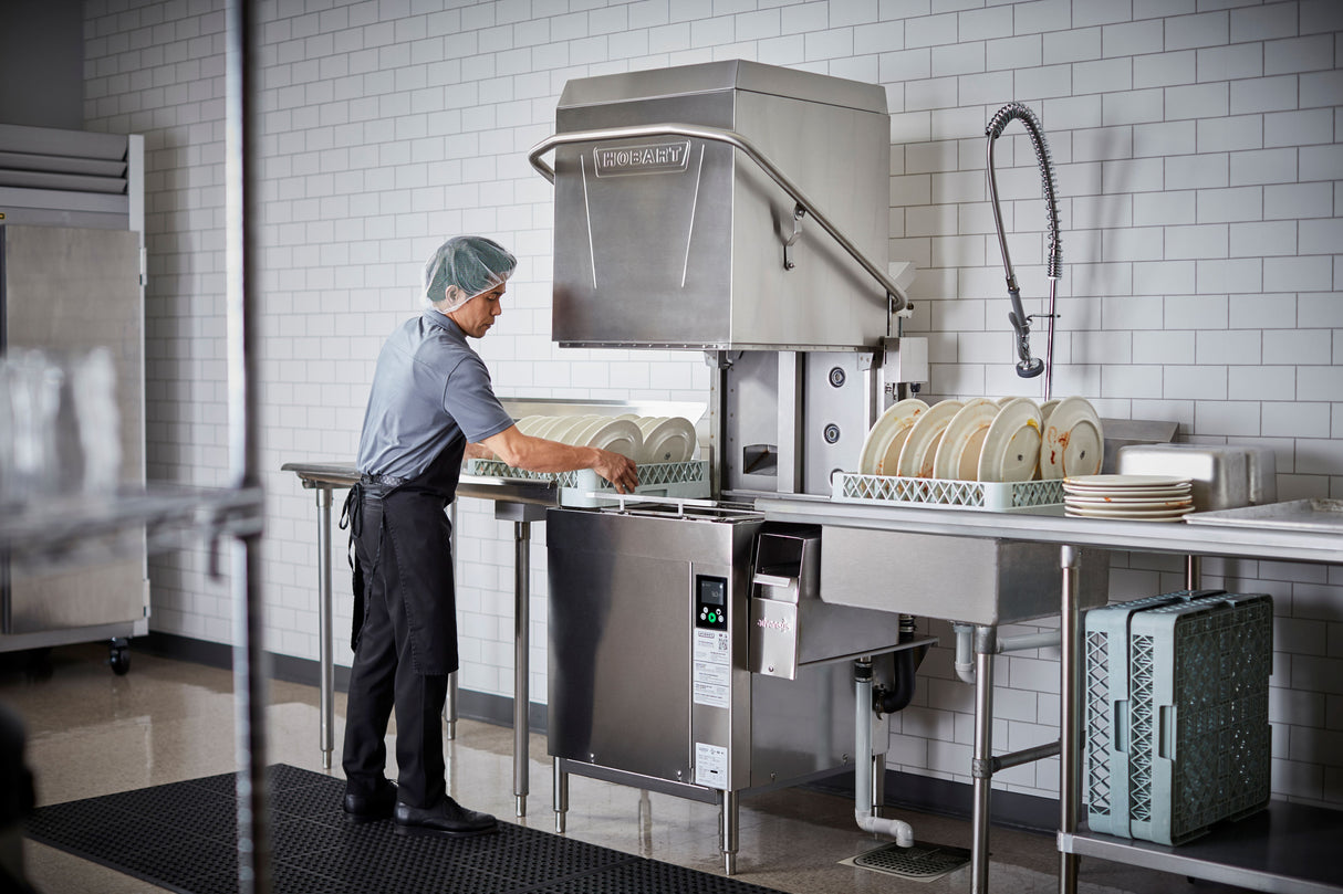A person wearing a hairnet and uniform loads dishes into a ventless dishwashing machine in a commercial kitchen. Stacks of plates sit on metal racks, while white tiled walls and stainless steel surfaces emphasize the clean, organized environment.