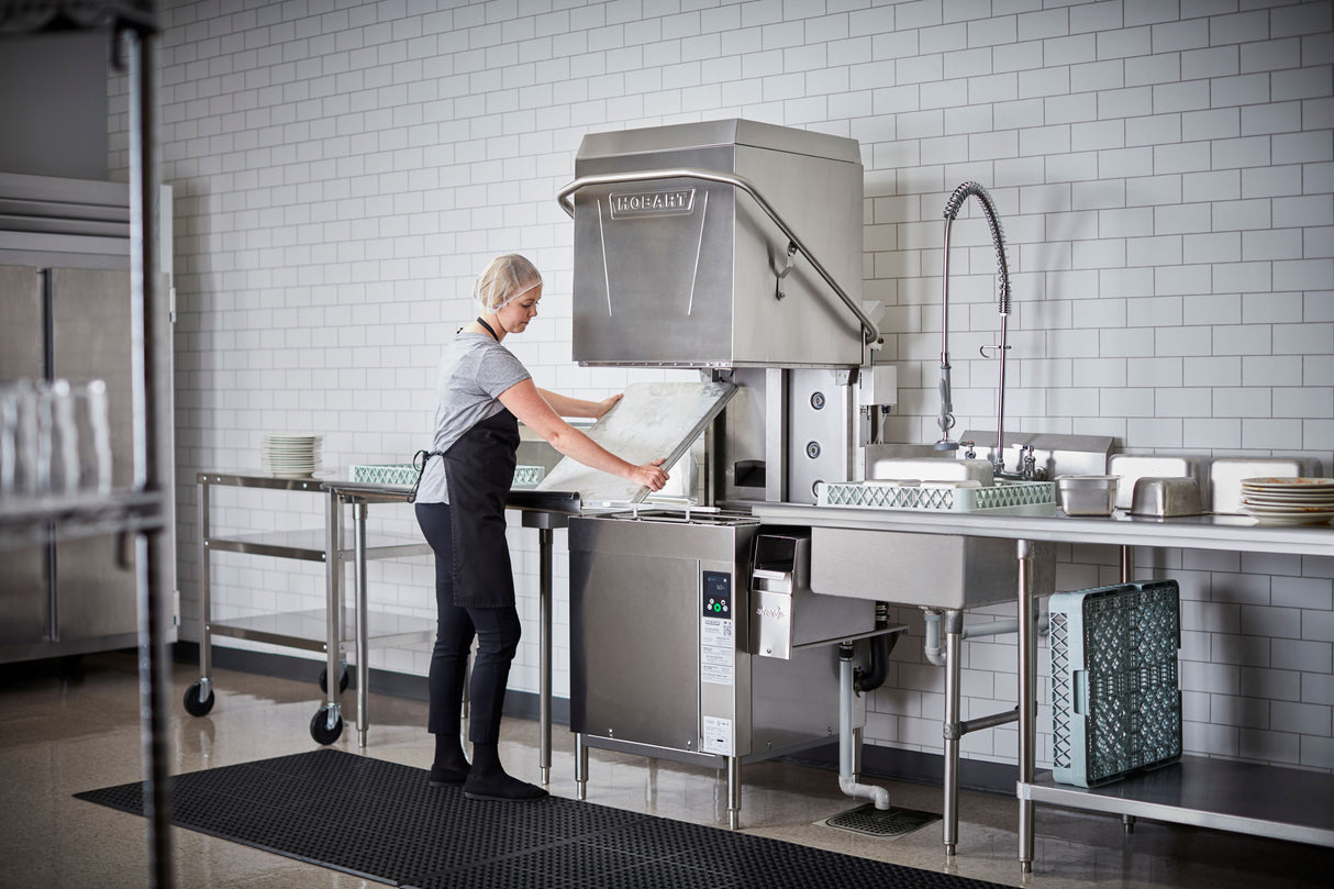 A person wearing a hairnet, apron, and gloves loads trays into a ventless dishwashing machine in a clean, tiled kitchen. Metal countertops hold stacks of dishes, while a sprayer hose and rack of washed, high temp sanitized dishes are nearby.