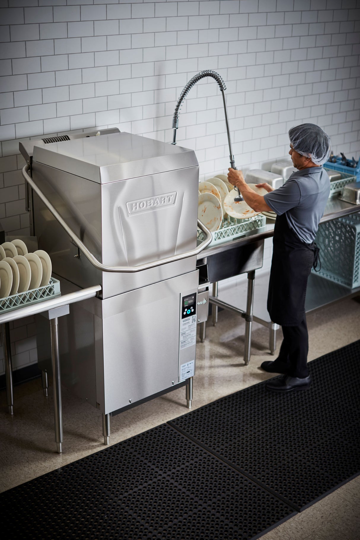 A person wearing a hairnet, apron, and gloves uses a flexible hose sprayer to rinse plates next to a high temp sanitizing, ventless dishwashing machine in a clean kitchen with white subway tile walls and black floor mats.