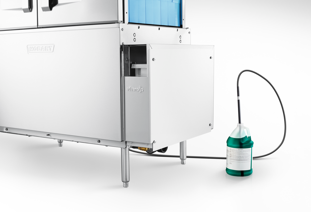 A conveyor dishwasher with a metal exterior stands on four legs. A tube connects it to a large green and white jug of cleaning solution on the floor. The scene, showcasing automatic soil removal, is brightly lit with a white background.