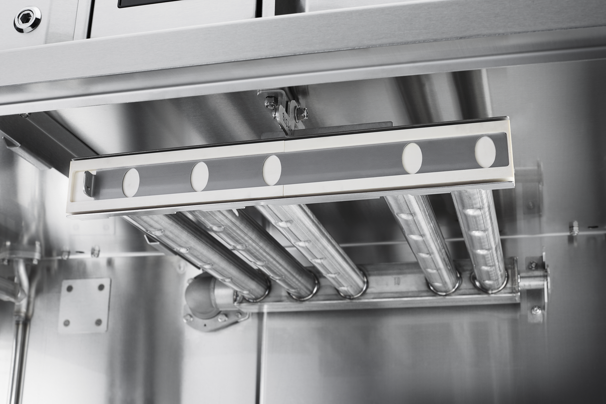 Close-up of an industrial stainless steel interior, likely part of a conveyor dishwasher, showing three parallel metal rods and a rectangular fixture. The clean, reflective surfaces suggest features like automatic soil removal in commercial kitchens or labs.