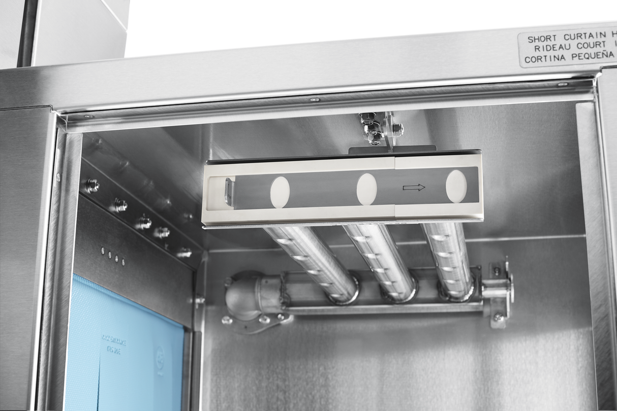 Close-up of a stainless steel conveyor dishwasher interior, featuring cylindrical metal pipes, wiring, and a box with circular windows and an arrow. A blue panel and small sign are visible—system supports automatic soil removal for efficient cleaning.