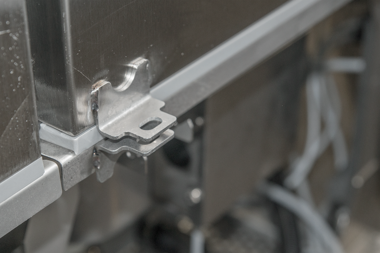 A close-up of a metallic bracket attached to the corner of a Hobart FT1000E flight type dish machine. The bracket is secured to the edge, with a rubber or plastic seal along the seam and wires and mechanical parts visible in the background.
