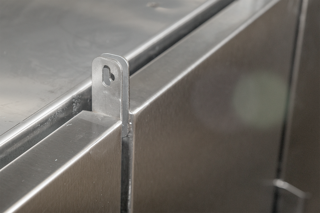 Close-up of a brushed metal latch or bracket attached to the edge of a stainless steel panel, possibly part of a ventless flight type dish machine. The bracket has a rounded top with a keyhole-shaped cutout. The surface is clean and reflective.