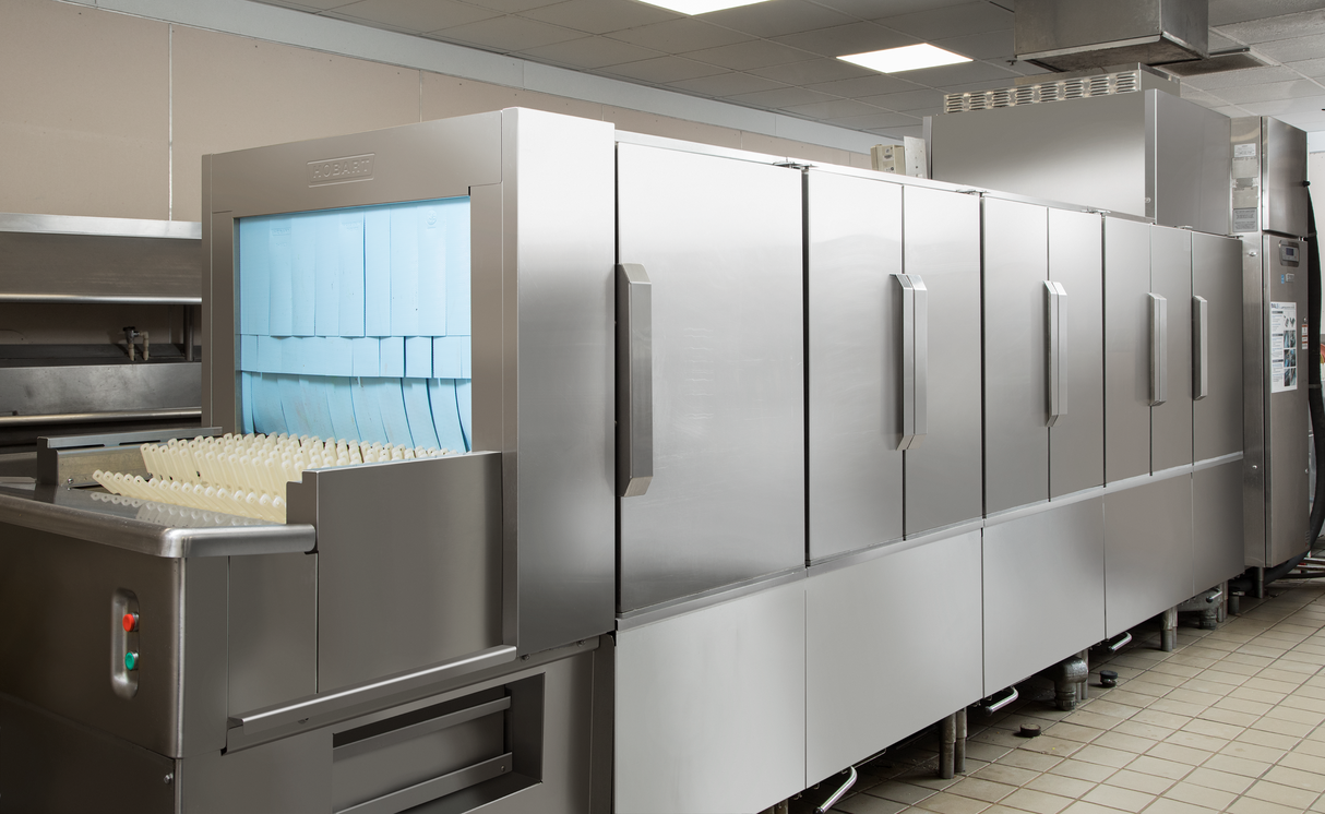 A large ventless flight type dish machine made of stainless steel stands in a commercial kitchen, with racks of blue trays and cups loaded inside. The machine features multiple compartments with handles and an automatic soil removal system.