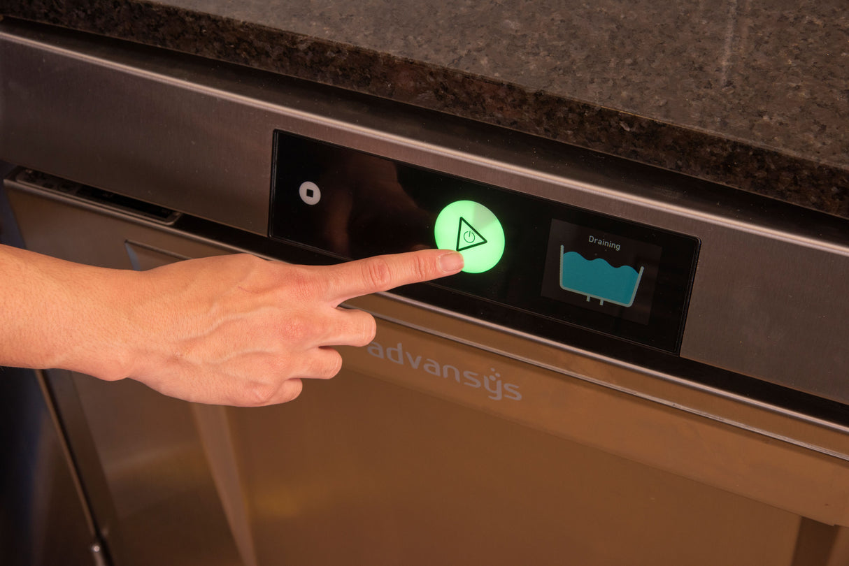 A hand presses a large, illuminated green play button on an undercounter dishwashers stainless steel panel. Next to it, a display shows a water tank draining icon. The countertop above is dark-colored stone. This model features high temperature sanitizing.