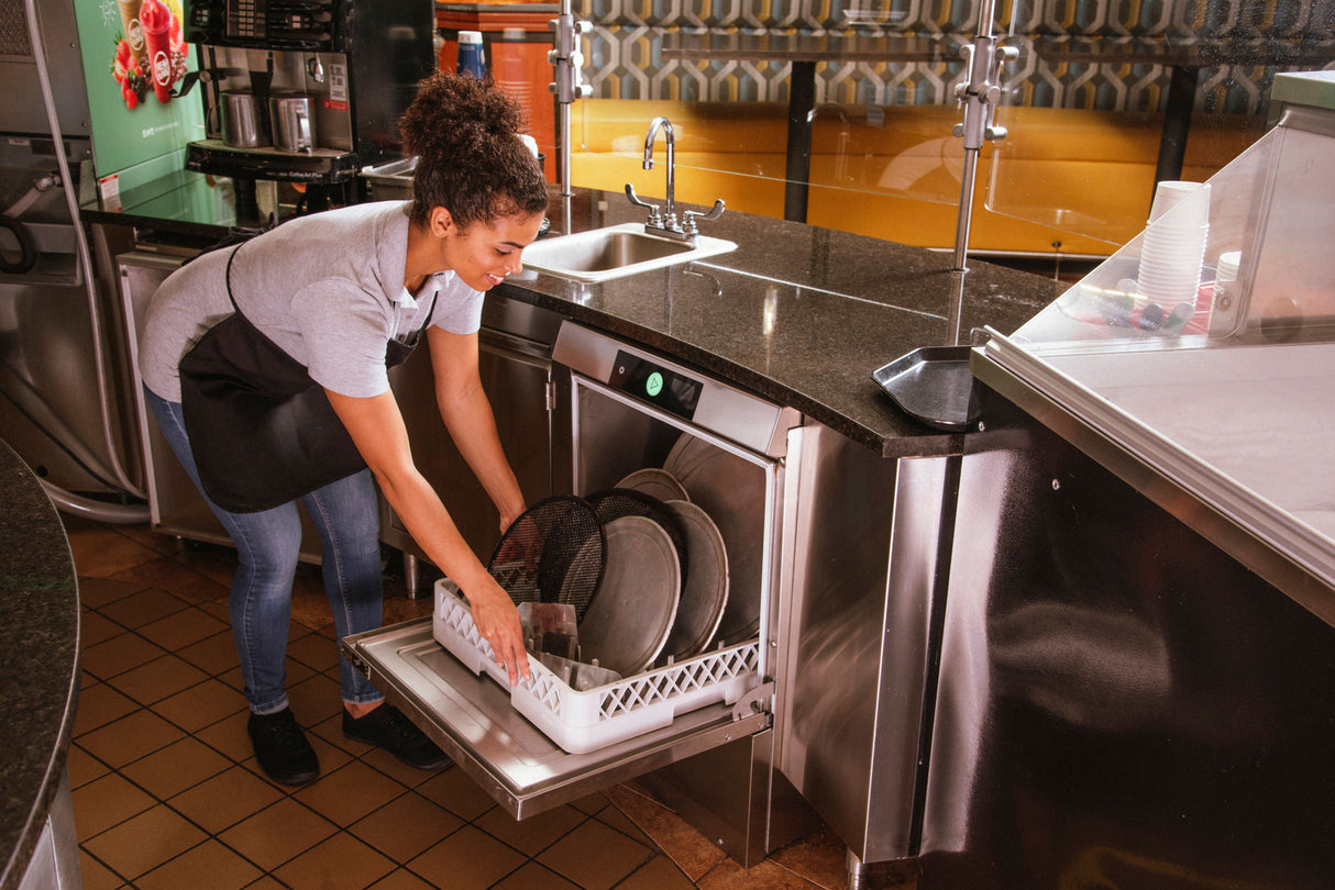 A woman wearing an apron loads metal trays into an ENERGY STAR undercounter dishwasher in a restaurant kitchen. She smiles as she works efficiently, surrounded by stainless steel counters, a sink, and other kitchen equipment.