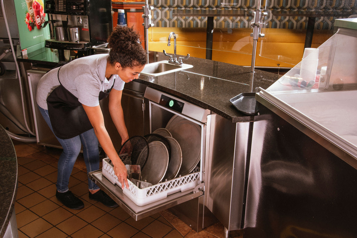 A woman wearing an apron loads metal trays and pizza screens into a Hobart LXn Dishwasher in a restaurant kitchen featuring high temperature sanitizing, black granite countertops, a stainless steel sink, tiled floor, and booth seating in the background.