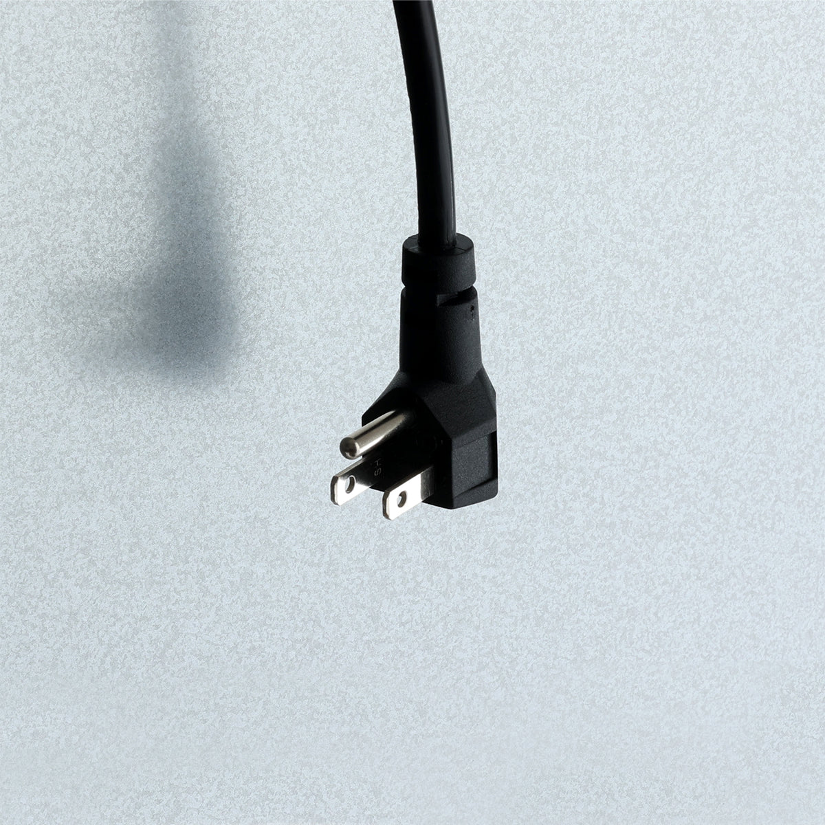 A black, three-prong electrical plug—perfect for a Reach-In Refrigerator—is shown against a light gray textured background. The plug is angled downward, with two flat prongs and one round grounding prong visible. The cord extends upward out of the frame.