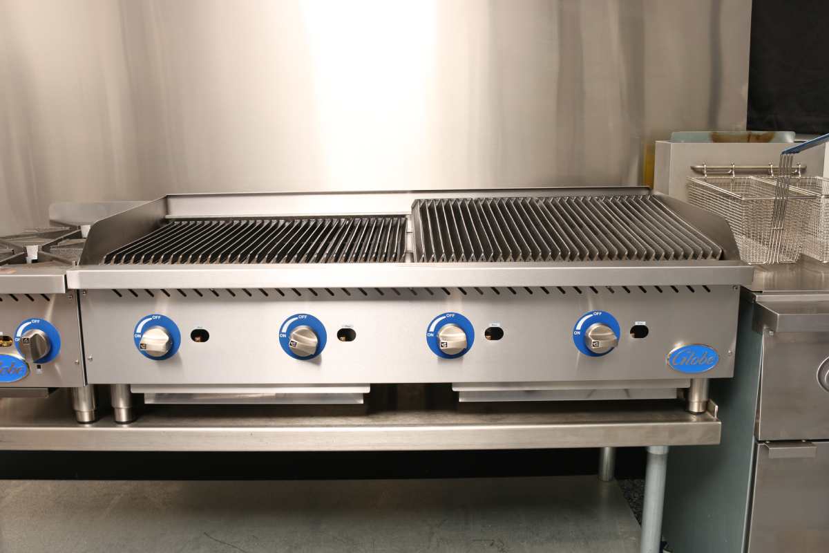 Commercial stainless steel gas charbroiler with five blue control knobs, dual ribbed grilling surfaces, and an adjacent deep fryer with metal baskets, set against a metallic wall in a professional kitchen environment.