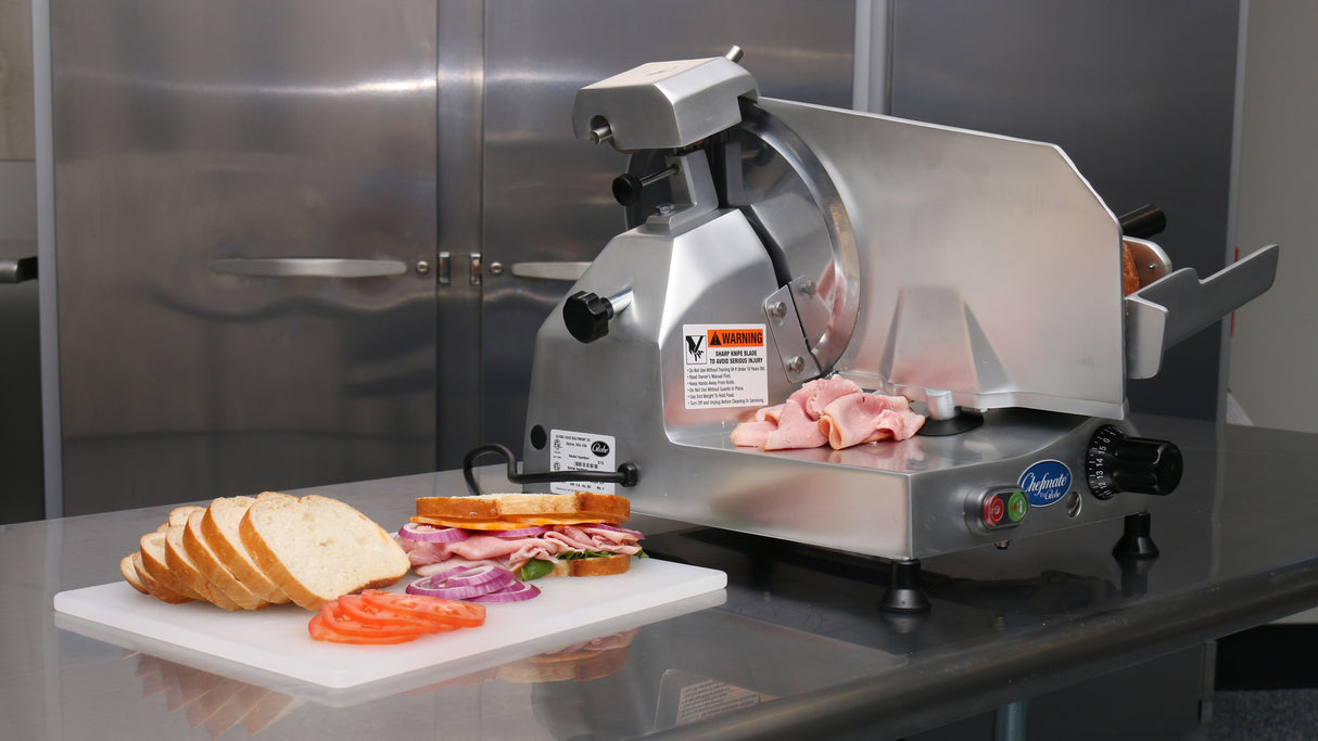 A food slicer sits on a stainless steel counter, slicing pink deli meat with its 12" knife. In front, a cutting board holds stacked sandwich ingredients: sliced bread, tomato, red onion, lettuce, meats, and cheeses. Metal cabinets are in the background.