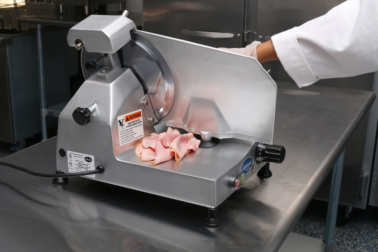 A person in a white coat uses a commercial 12" knife food slicer to cut pink deli meat in a stainless steel kitchen. The slicer has warning labels and controls, and thin slices of meat are stacked neatly on the machine’s tray.