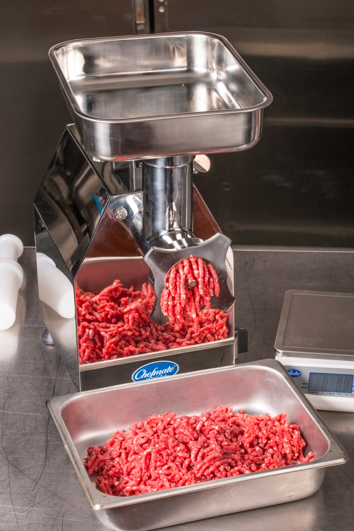 A Chefmate meat grinder with a #12 head size extrudes ground beef into a metal tray. A digital scale and white plastic attachments sit nearby on a stainless steel counter in the clean, industrial space.