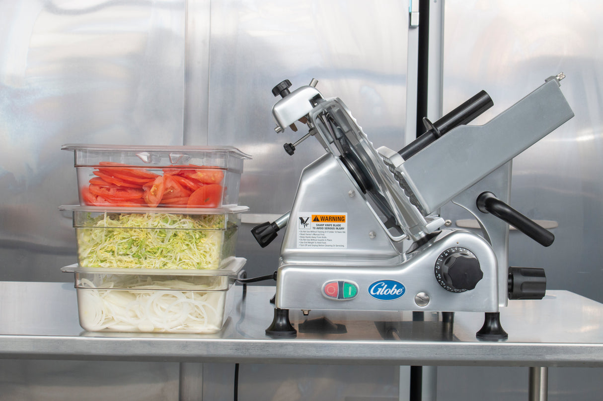 A Globe G12 12" manual slicer sits on a metal counter next to three stacked clear containers filled with sliced tomatoes, shredded lettuce, and sliced onions. The reflective metal surface in the background suggests an industrial kitchen setting.