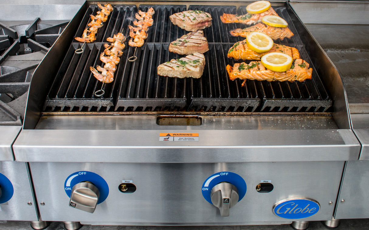A gas charbroiler with heavy duty cast-iron grates sizzles with shrimp skewers, seasoned fish fillets, and two salmon fillets topped with lemon slices. The stainless steel Globe grill features blue-accented knobs marked "ON" and "OFF.