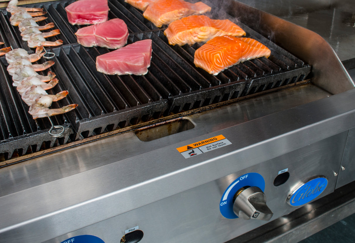 Close-up of a gas charbroiler with raw shrimp skewers, pink tuna steaks, and orange salmon fillets sizzling on heavy duty cast-iron grates. Blue temperature control knobs and a warning label are visible; steam rises from the fresh seafood.