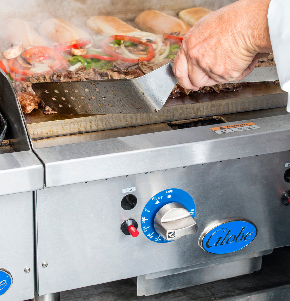 A hand holds a metal spatula, grilling chopped meat and vegetables on a gas countertop griddle. Steam rises as sandwich buns toast in the background. The stainless steel front features thermostatic controls and the Globe brand label.