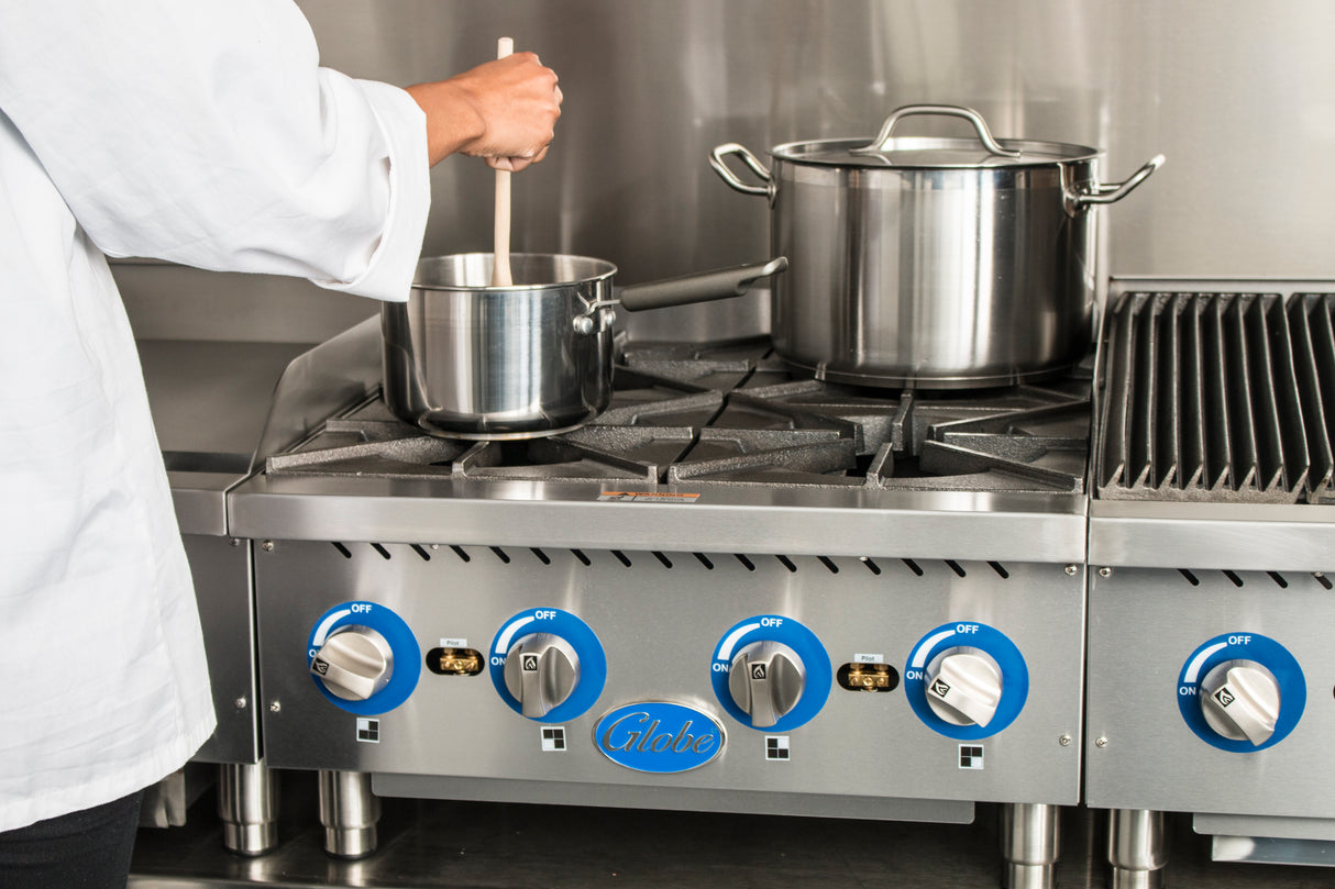 A person in a white chef coat stirs a small pot on a commercial gas countertop hotplate with blue knobs; another larger covered pot and grill plate sit on the Globe GHP24G with cast iron burners in a professional kitchen setting.