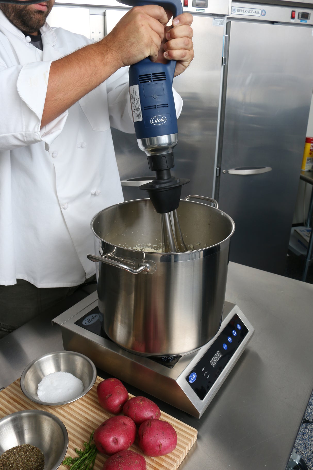 A chef in a white coat uses an Immersion Blender with a removable stainless steel shaft to blend food in a metal pot on an induction cooktop. On the counter are red potatoes, rosemary, salt, and pepper. Stainless steel refrigerators are in the background.