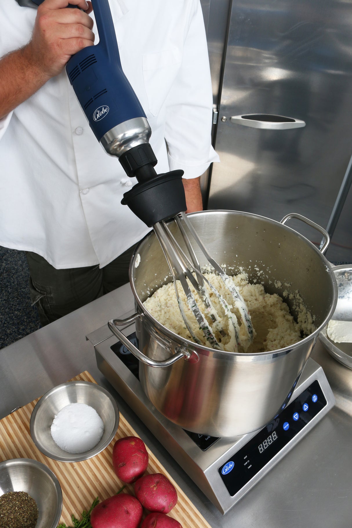 A chef in a white coat uses a variable speed Immersion Blender with a removable stainless steel shaft to mash potatoes in a pot on an induction cooktop. Red potatoes, salt, pepper, and fresh herbs rest on a striped mat nearby. Stainless steel appliances gleam behind.