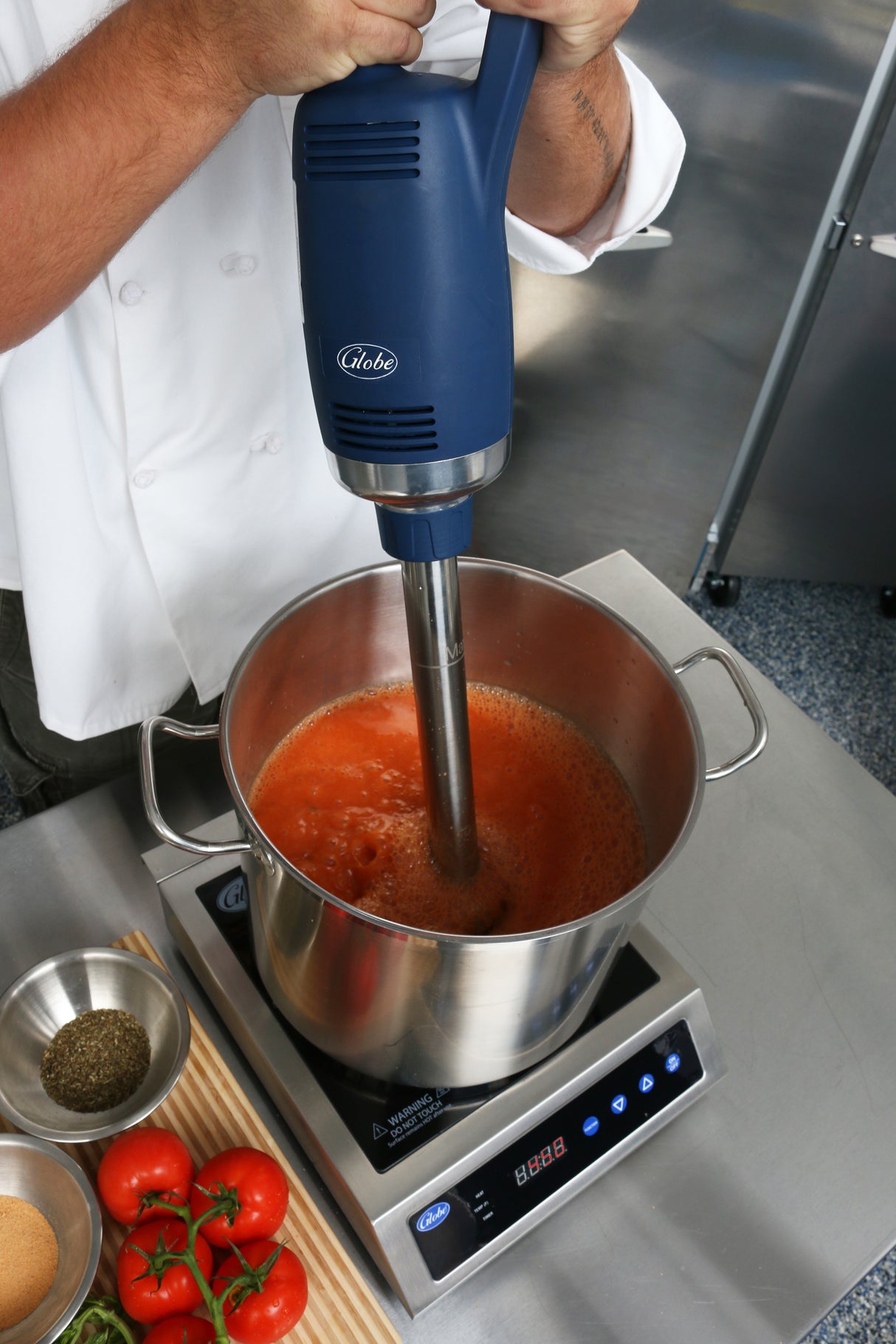 A chef uses a variable speed Immersion Blender with a 14" stainless steel shaft to puree red tomato soup in a stainless steel pot on an induction cooktop. Fresh tomatoes, herbs, and spices are arranged nearby; only the chef's arms and hands are visible.