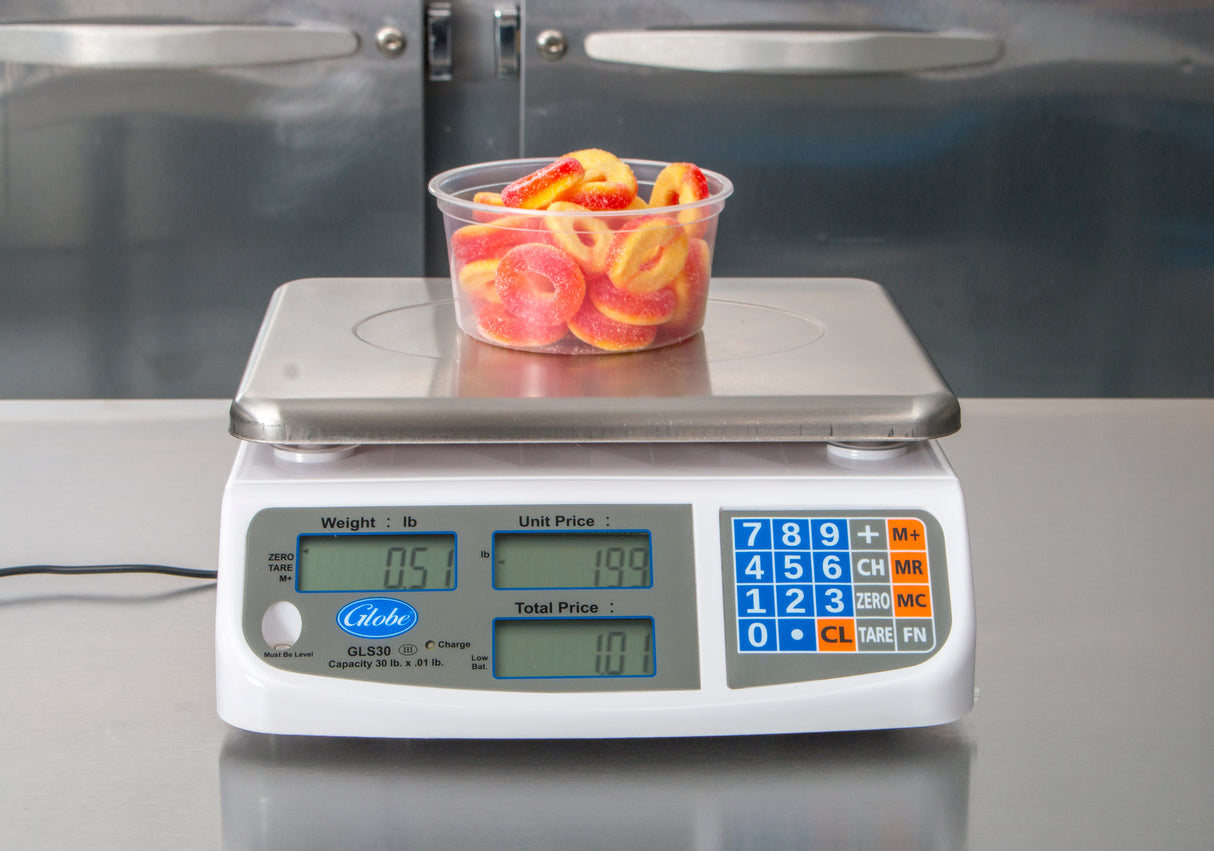 A clear plastic tub filled with gummy peach ring candies sits on a price computing scale. The scale, featuring a dual LCD display, shows 0.51 lbs at $1.99 per pound for a total of $1.01. The background is a metallic kitchen surface.