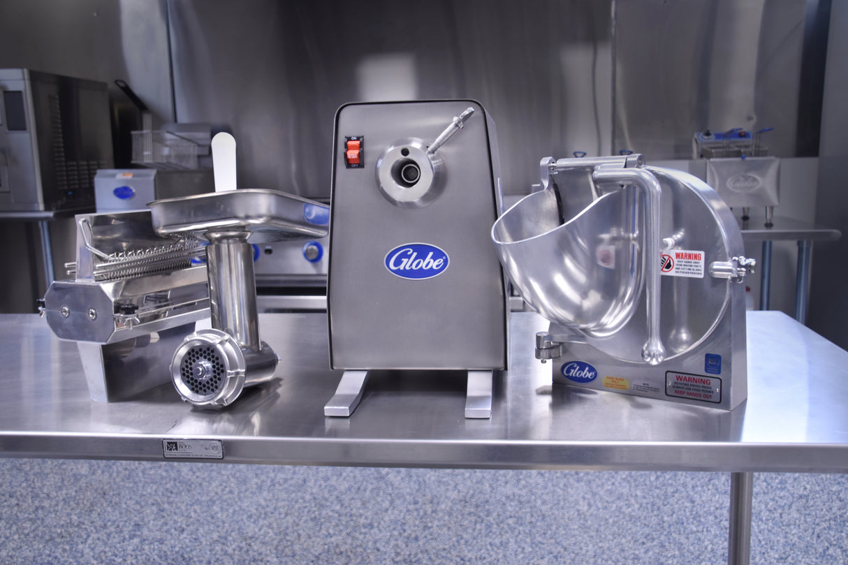 A stainless steel commercial meat grinder with 1 HP Power Drive Unit and food processor sit on a metal counter in an industrial kitchen. Both machines feature “Globe” logos, large attachments, and durable stainless steel construction.