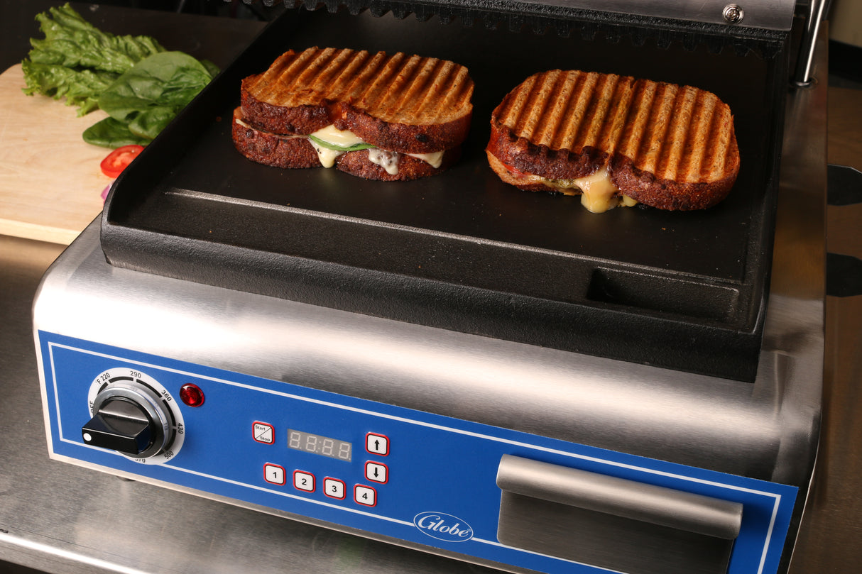 Two grilled sandwiches with visible grill marks cook on a countertop electric grill. Melted cheese oozes from the sides, while fresh greens and sliced tomatoes rest on a cutting board behind the stainless steel unit with digital controls.