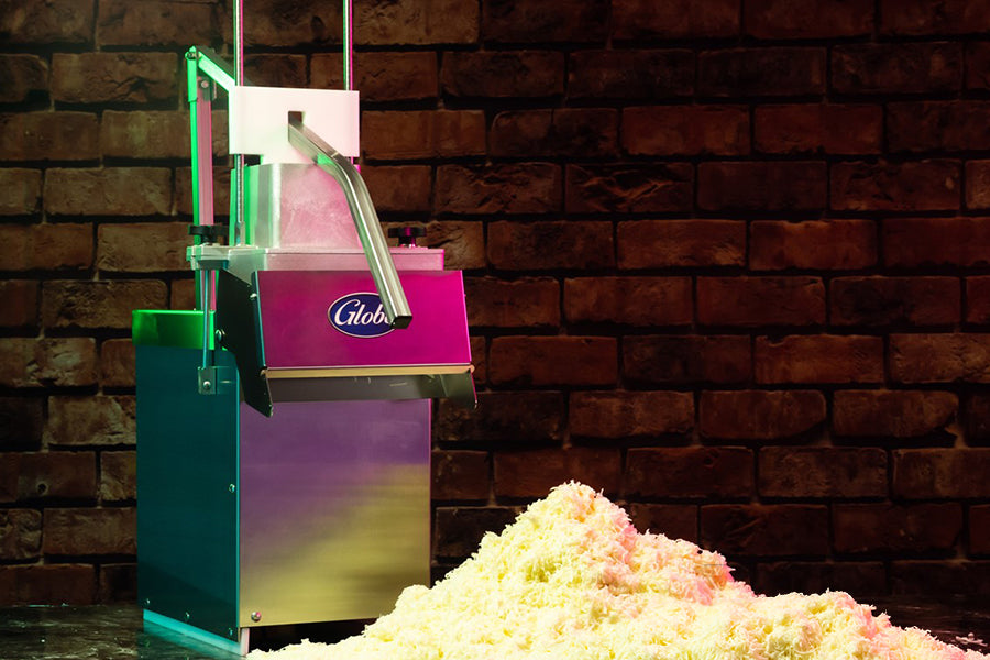 A shiny industrial cheese shredder with continuous feed sits on a floor against a brick wall, dispensing a large pile of freshly grated cheese beneath its chute. The high volume Globe machine features a sleek metallic finish and prominent branding.